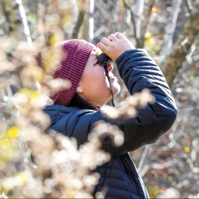 Photograph of Kristine Meader ’21 among the trees on campus, wearing a down jacket and red knit cap, using binoculars to look for birds