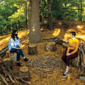 Valeria Yang ’21 and Bryn van Dommelen ’22 sit on tree stumps in the Sitting Circle in golden early fall light