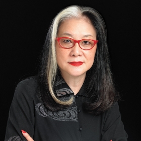 A photo of Mee See Loong '72, wearing black, with red-framed eyeglasses and a dramatic red ring on her hand.