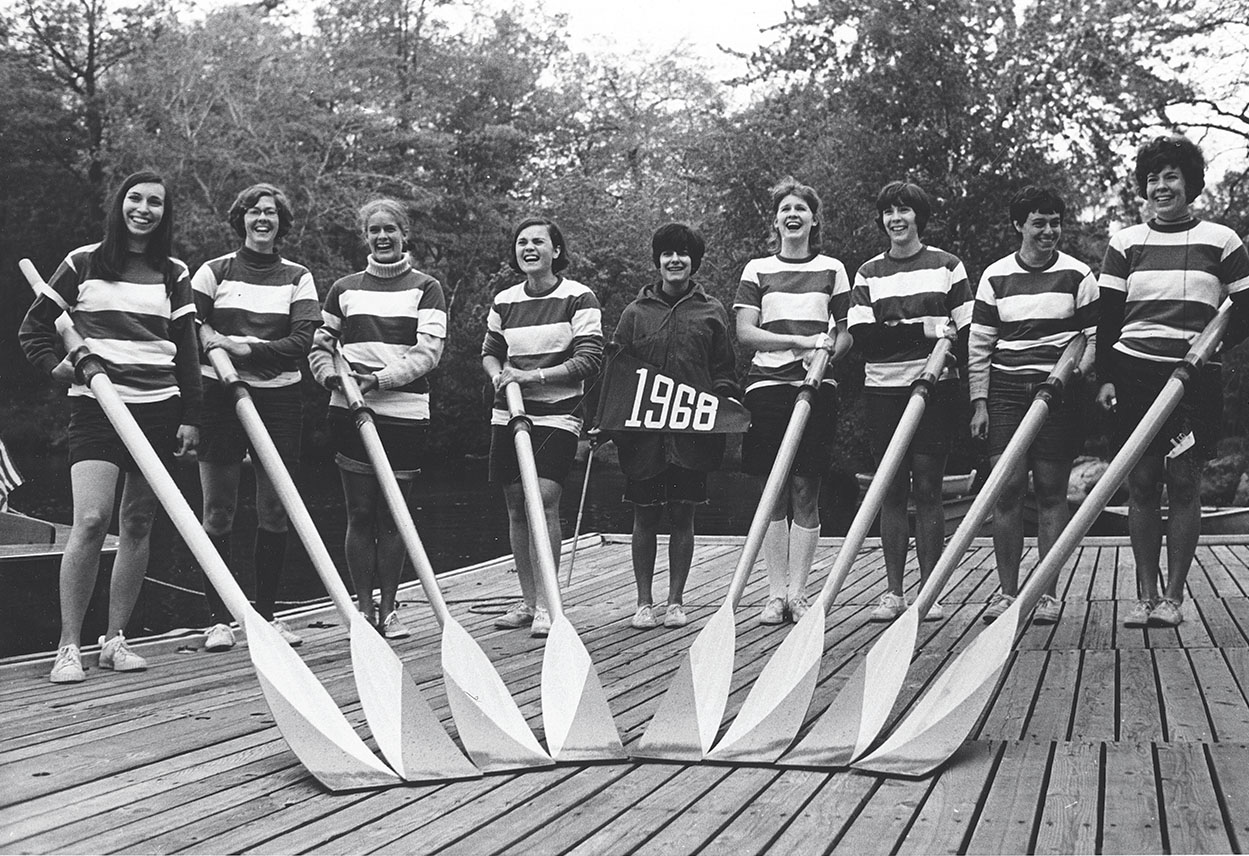 A black and white photo of students posing on a dock with their oars in front of them