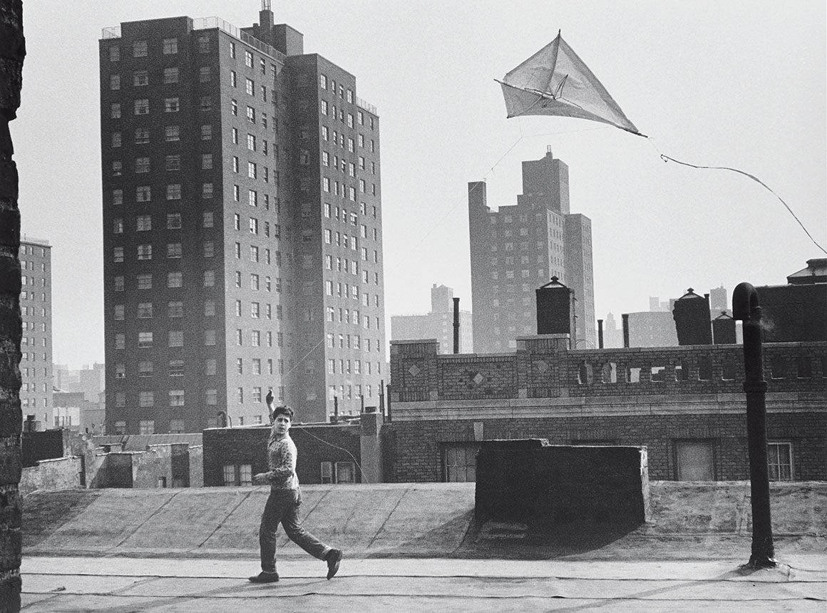 A black-and-white photo of a boy flying a kite on the rooftop in a cityscape