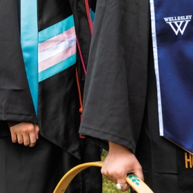 A photo shows a close up of a trans flag stole worn by a graduating senior. In the background another student wears a Pride fag stole.