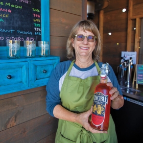 A photo shows Kelly Miller McCune holding a large bottle of her product at the Runcible Cidery in Oregon.