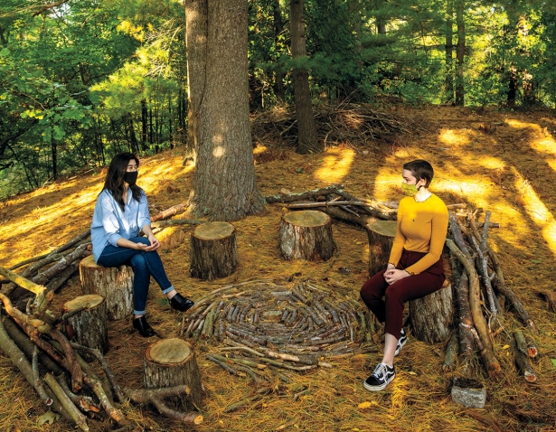 Valeria Yang ’21 and Bryn van Dommelen ’22 sit on tree stumps in the Sitting Circle in golden early fall light