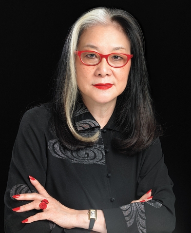 A photo of Mee See Loong '72, wearing black, with red-framed eyeglasses and a dramatic red ring on her hand.