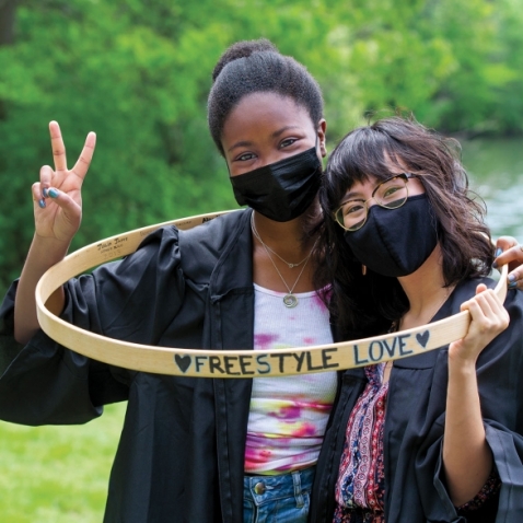 Tendayi Peyton ’21 and Izabelle Fernandez ’21 pose inside a hoop reading "Freestyle Love." Both are wearing masks and graduation robes. 