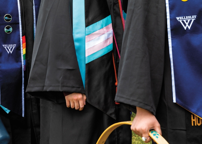 A photo shows a close up of a trans flag stole worn by a graduating senior. In the background another student wears a Pride fag stole.