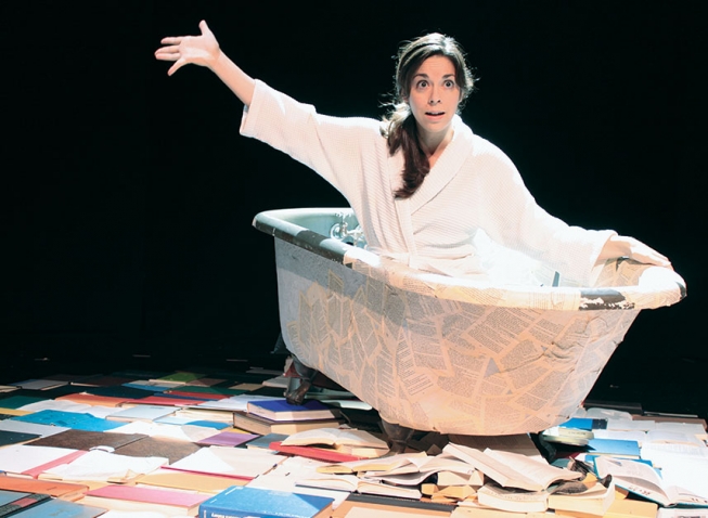 Marta Rainer '98 sits in a bathtub on stage surrounded by books as part of her one-woman show