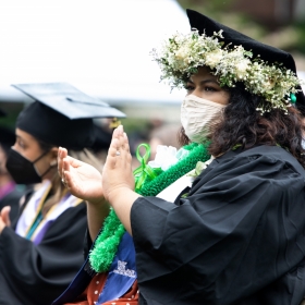 A student with a white wreath of flowers around her tam applauds at commencement
