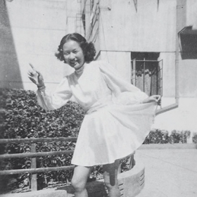 A young woman poses for the camera pointing with one hand while holding up a corner of her skirt in a kind of curtsy with the other
