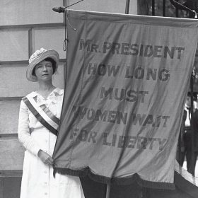 A suffragist holds a banner reading "Mr. PRESIDENT HOW LONG MUST WOMEN WAIT FOR LIBERTY"
