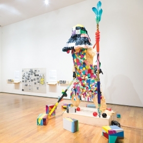 Survival Robot (2020), a two-story tall humanoid figure holding two colorful staffs and wearing a shift made of colorful, pieced-together shapes, surrounded by smaller colorful boxes