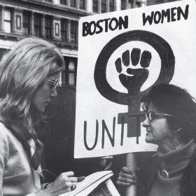 Lynn Sherr interviewing a participant holding a sign reading "UNITE" with a female symbol and a fist during a women's rights march in 1970